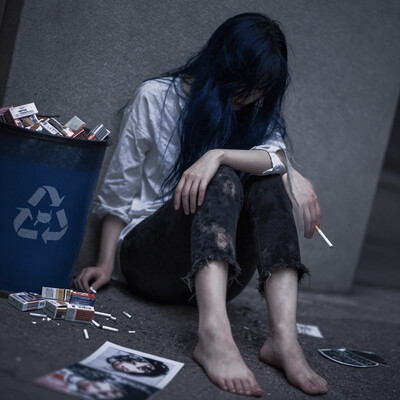 Dutch tilt, Low-angle shot, 35mm wide-angle lens with mild barrel distortion, deep f/5.6 depth of field. Dimly lit scene with muted grays and blues. A young woman with long, dark blue hair and a white shirt sits against a grey wall, her face hidden behind her hair and left arm, holding a cigarette. Her black pants are stained and her feet are bare, with a few cigarette butts scattered on the floor. A trash can overflows with cigarette packs, a blue recycling bin has a cat print on it, and a few scattered photographs show a person with a similar face. The artistic style is of manga.