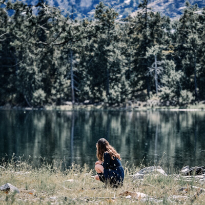 masterpiece, newest, absurdres, incrediblvy absurdres, best quality, amazing quality, very aesthetic, 1girl, squatting on grass, mountain lake, clear sky, rocky shoreline, reflections on water, tall pine trees, natural lighting granbluefantasysyleil